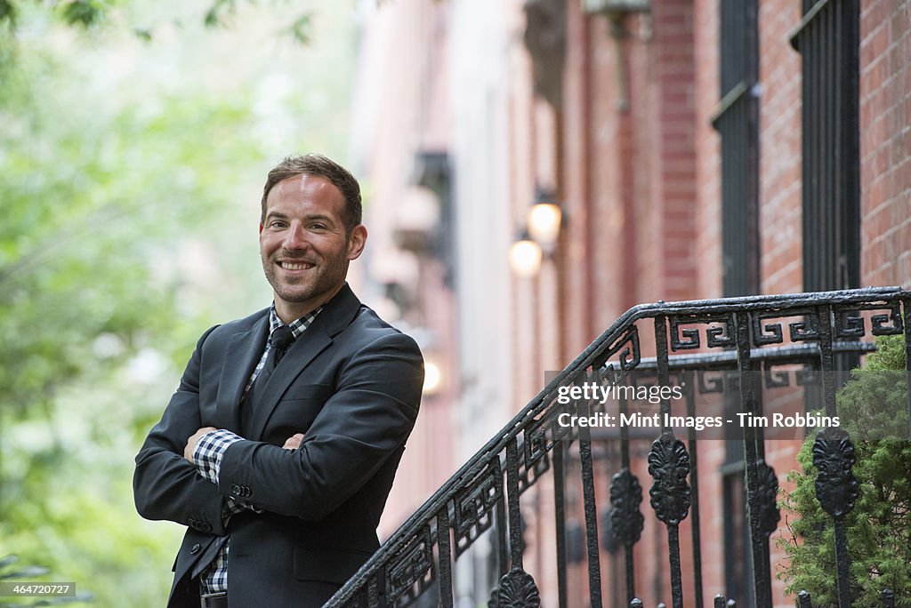 Business people out and about in the city. A man in a suit on the steps of a brownstone building. Arms folded.