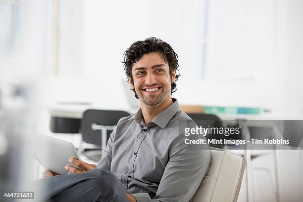 an office in the city. business. a man seated smiling and holding a digital tablet. - asian man sitting at desk ストックフォトと画像
