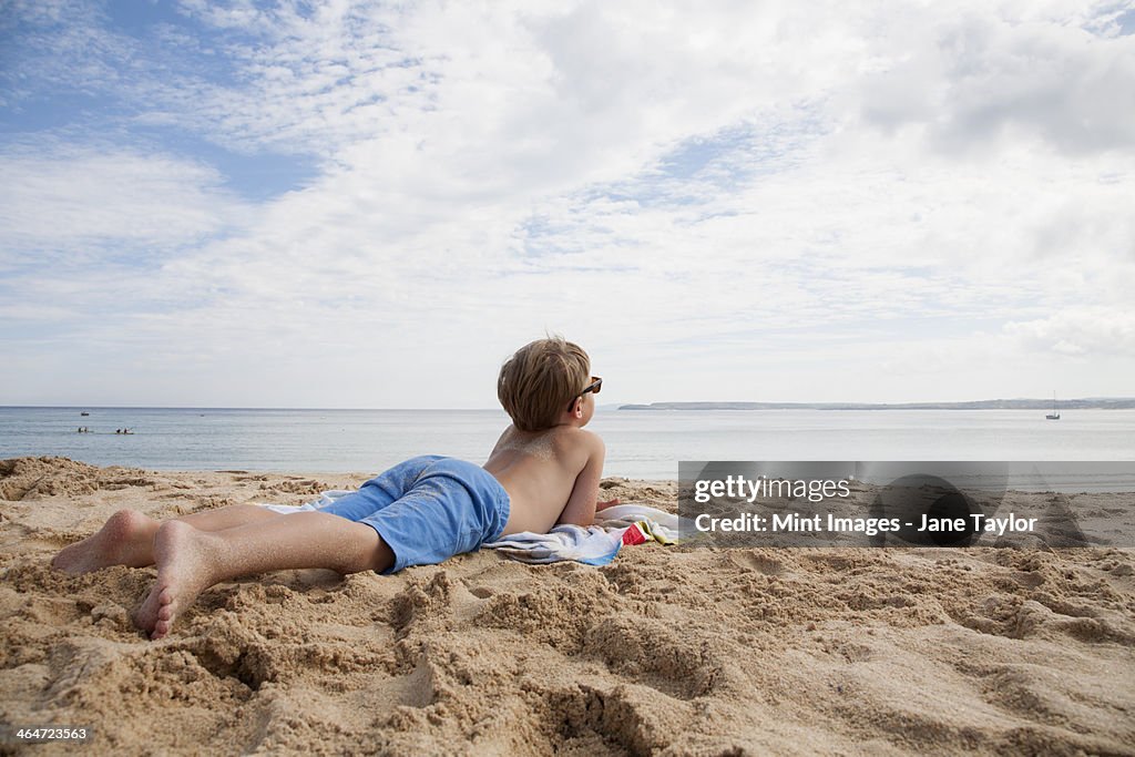 A boy lying on his front on the sand looking out to sea.