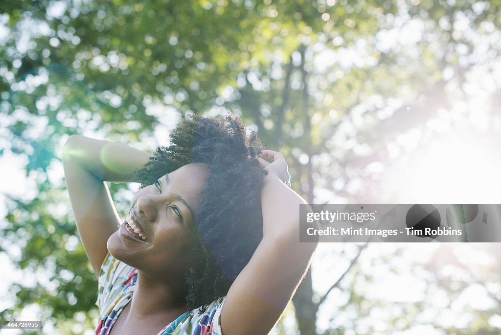 A young woman in a flowered summer dress with her hands behind her head, smiling and looking up.