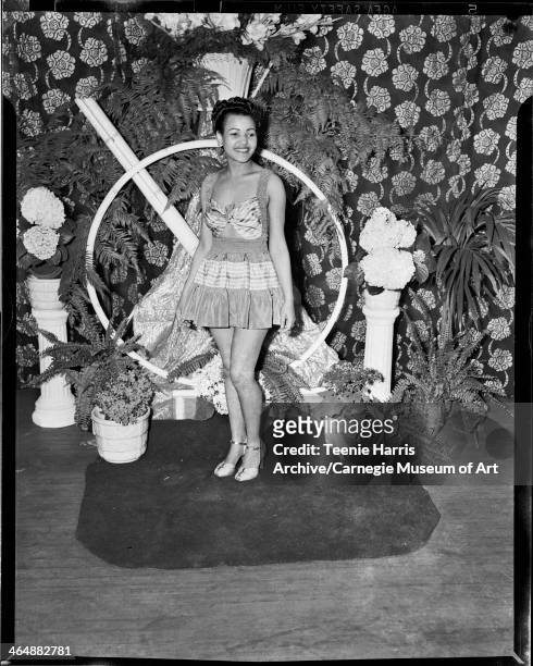 Woman wearing short sundress, standing against backdrop of floral curtain, ferns, and hydrangeas, for Beauty Shop Owners' Fashion Review in Schenley...