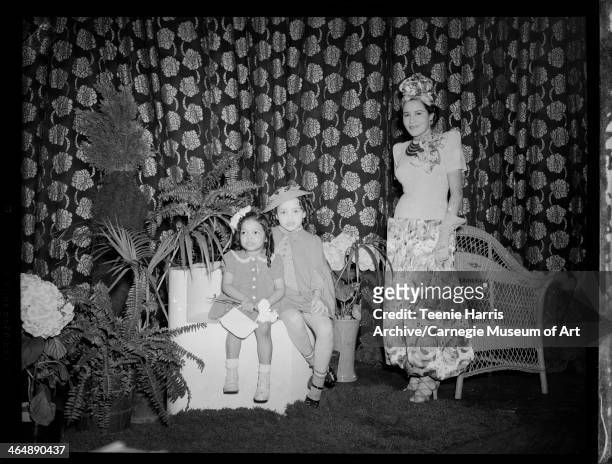 Woman standing beside wicker armchair and two seated girls, posed in front of floral curtain, feathers, ferns, and hydrangeas, for Beauty Shop...