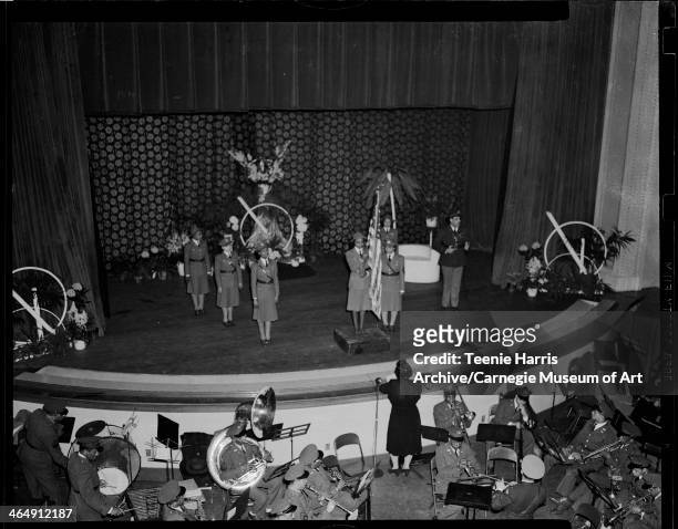 Color guard presenting flag on stage with floral curtain, feathered decoration, hydrangeas, and abstract metal decorations, with orchestra in pit,...