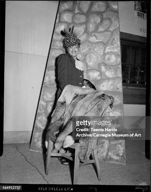 Woman wearing hat with two feathers, standing behind chair covered with furs in front of painted stone backdrop, for Beauty Shop Owners' Fashion...