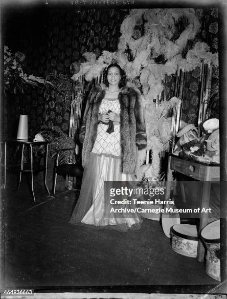 Leona Worthington wearing evening dress with diamond patterned tunic and sheer skirt under fur coat, on stage with floral curtain, feathers, and hat...