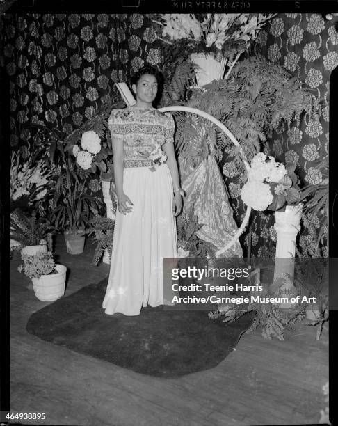 Woman wearing floral short-sleeved top and floor length skirt with corsage at waist, on stage with floral curtain, hydrangeas in pots, and abstract...