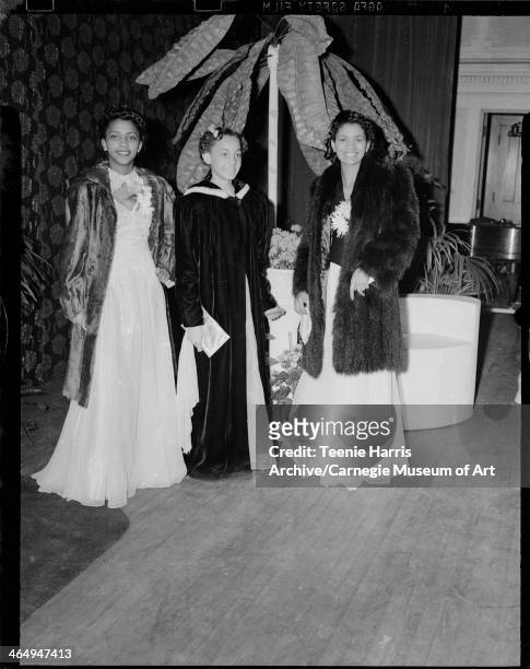Group portrait of three women, two wearing fur coats, and one wearing floor-length coat with light colored fur trimmed hood, posed on stage with...