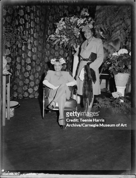 Two women, one seated in armchair, modeling day dresses, hats, and fur stole, on stage with floral curtain and potted hydrangeas, for the Beauty Shop...