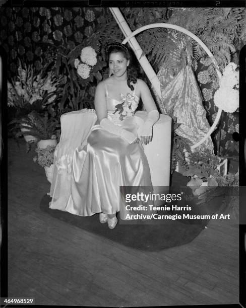 Woman wearing satin gown and corsage on bodice, posed in interior with floral curtain, ferns, and hydrangeas, for Beauty Shop Owners' Fashion Review...