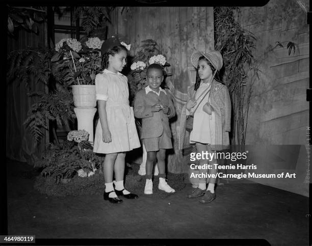 Group portrait of boy, and two girls, one wearing plaid coat and bonnet, posed on stage surrounded by ferns, hydrangeas, and bamboo, for Beauty Shop...