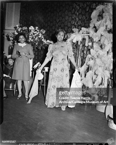 Woman wearing long floral dress standing beside woman wearing suit, against backdrop of floral curtain, feathered decoration, and hydrangeas, for...
