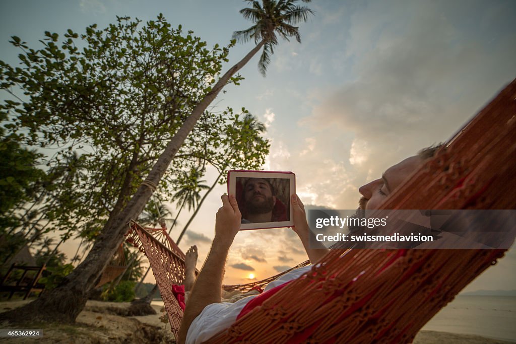 Man resting on hammock on tropical beach-Using digital tablet