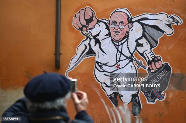 Man takes a picture of a street art mural showing Pope Francis as a superman, flying through the air with his white papal cloak billowing out behind...