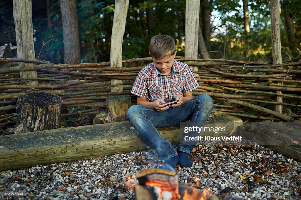 Boy playing game on phone, sitting by fire outside