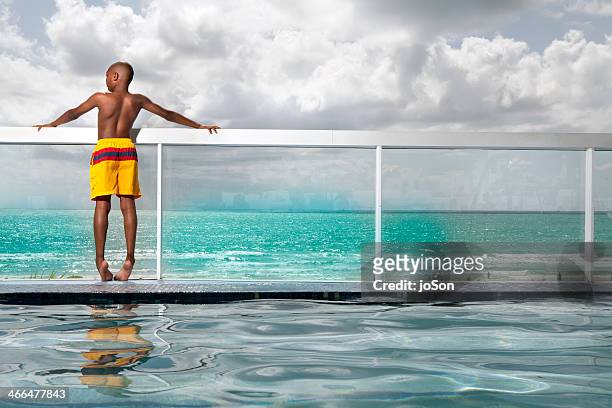 boy standing at the pool side looking at ocean - bermuda shorts stock pictures, royalty-free photos & images