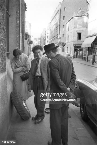 Swiss artist Alberto Giacometti stands on a sidewalk and talks with a man as, beside them, another man changes the tire on a parked car, Paris,...