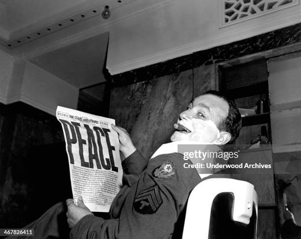 Sergeant interrupts his shave in a barber shop and holds up the latest copy of The Stars And Stripes announcing the surrender of Japan with the...