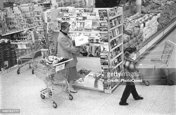 Woman reads a magazine as a small boy walks past in a large supermarket, Maryland, 1957. Paper-bound pocket books and magazines ahd recently been...