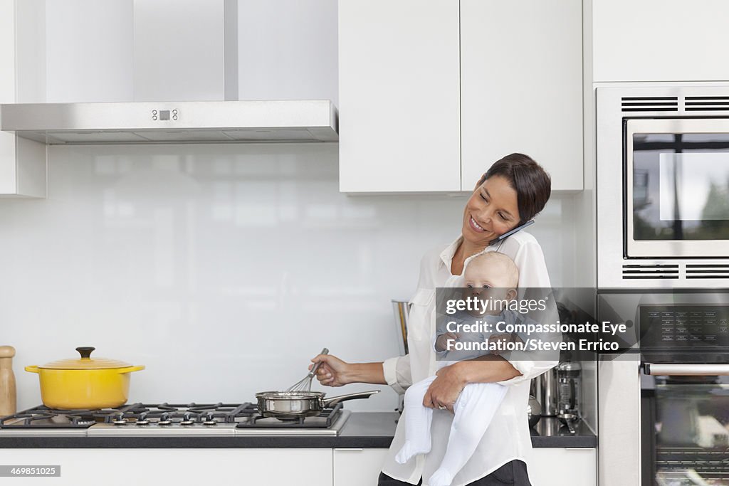 Mother holding baby in kitchen using mobile phone