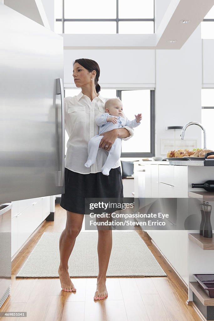 Mother holding baby in kitchen opening fridge