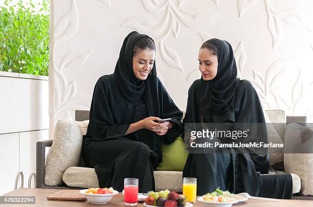 two emirati women in abaya texting on cellphone at lunch - 阿拉伯 個照片及圖片檔