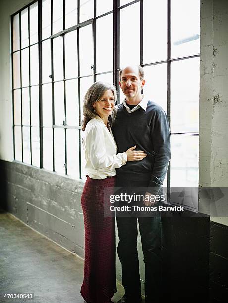 mature couple embracing by windows in loft space - mann 50 style stock-fotos und bilder