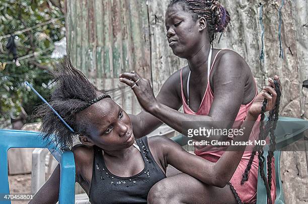 San Pedro de Macoris Dominican Republic slums, local beauty parlor. One woman with keloids on her arm combs out hair of another seated woman who...