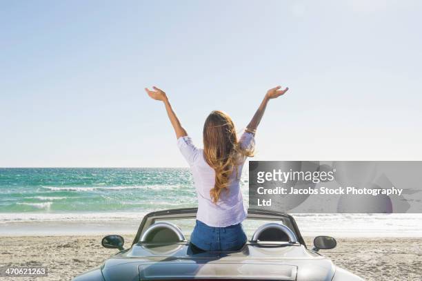 hispanic woman sitting on convertible at beach - braccia alzate foto e immagini stock