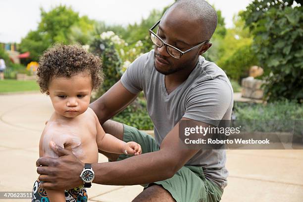 father putting sunscreen on toddler son in park - baby suncream stock pictures, royalty-free photos & images