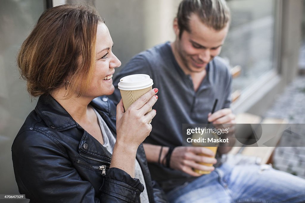 Happy woman drinking coffee in disposable cup with man on bench at sidewalk