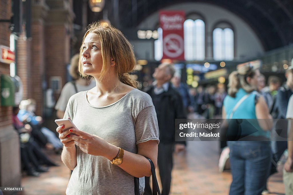 Mid adult woman with mobile phone looking up while standing on railway station