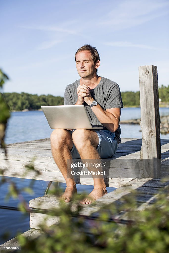 Mature man praying while sitting with laptop on pier