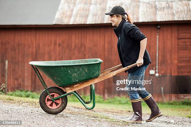 side view of female farmer pushing wheelbarrow on rural road - schubkarre stock-fotos und bilder