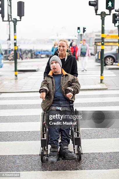 caretaker pushing disabled man on wheelchair while crossing road - rollstuhl schieben stock-fotos und bilder