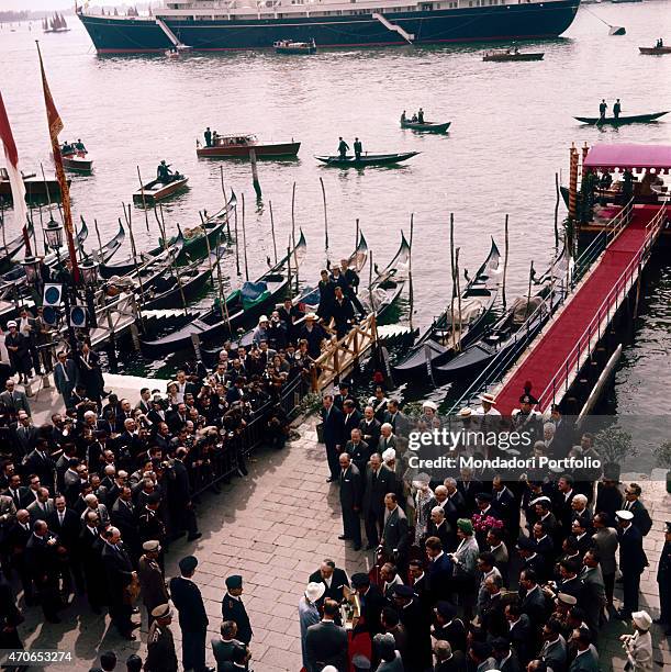 "Queen Elizabeth and Prince Philip, just disembarked the Yacht Britannia, are standing in the embankment of San Marco, surrounded by a big crowd; the...