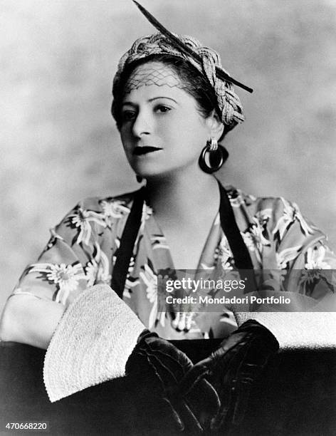 "Helena Rubinstein portrayed on a photo studio with a fancy American dress; the Polish born American entrepreneur wears a braided rope crown, a pair...