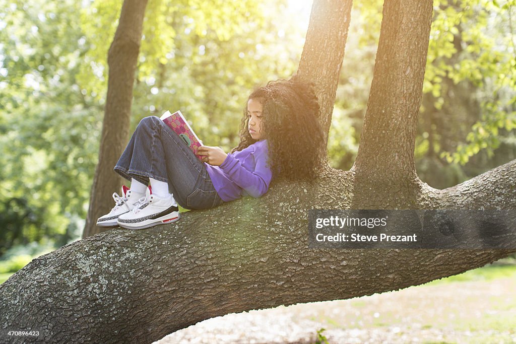 Young girl lying on tree branch reading book