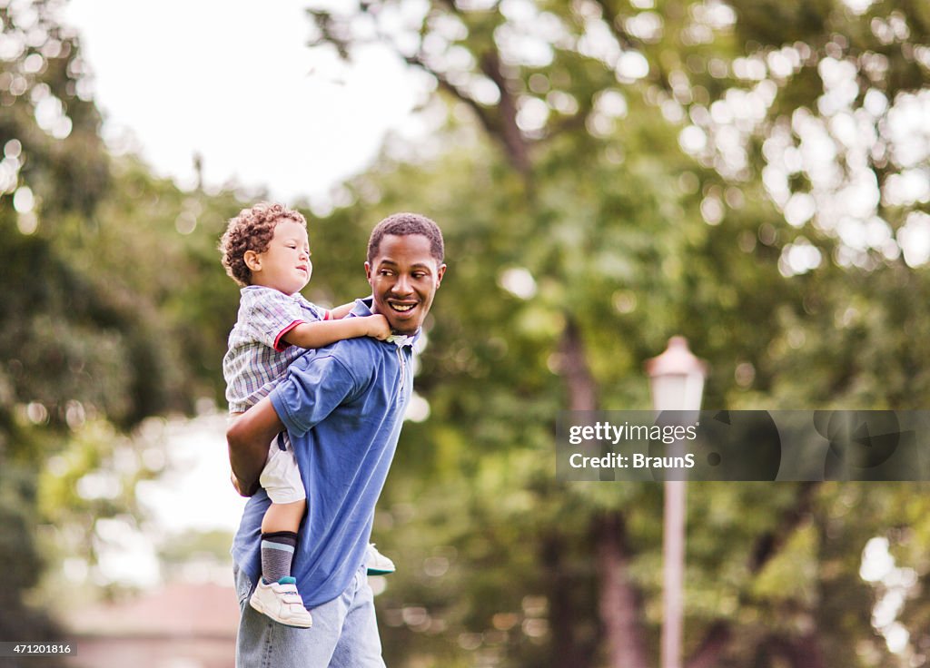 Playful African American father piggybacking his son outdoors.