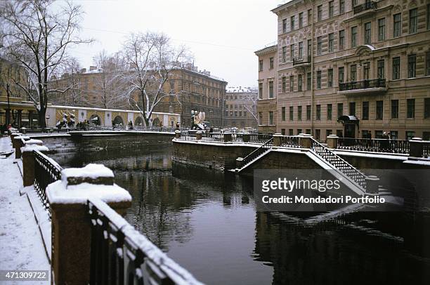Griboyedov canal after a snowfall, with the bronze griffins of the Bank Bridge, on the background, slightly covered with snow; on the left it is...