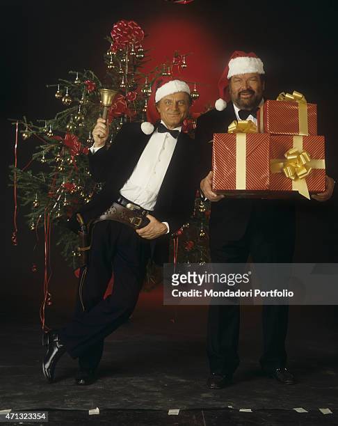Italian actor Terence Hill with a Santa Claus hat posing shoulder to shoulder with Italian actor Bud Spencer holding some gifts. They're the main...