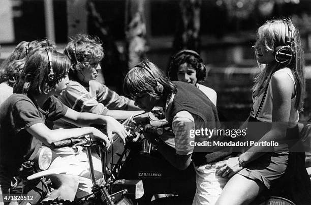 Some teenagers listening music in the headphones seated on their scooters. A boy and a girl sitting on the saddle of a Fantic Motor Caballero. Italy,...
