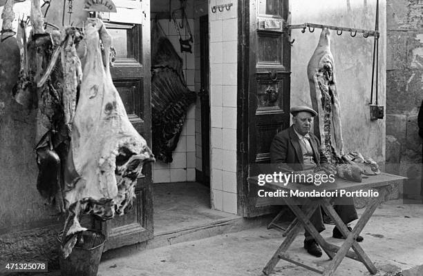 An Italian butcher sitting at the entrance of his shop between the carcasses of a veal and a pig. Agrigento, 1950s