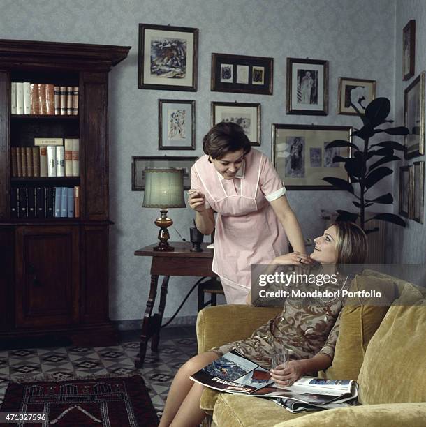 Housemaid smiling with the landlady seated on the sofa. Italy, 1960s