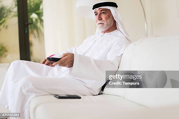 middle eastern senior watching tv at home dressed in dishdasha. - old arab man stock pictures, royalty-free photos & images