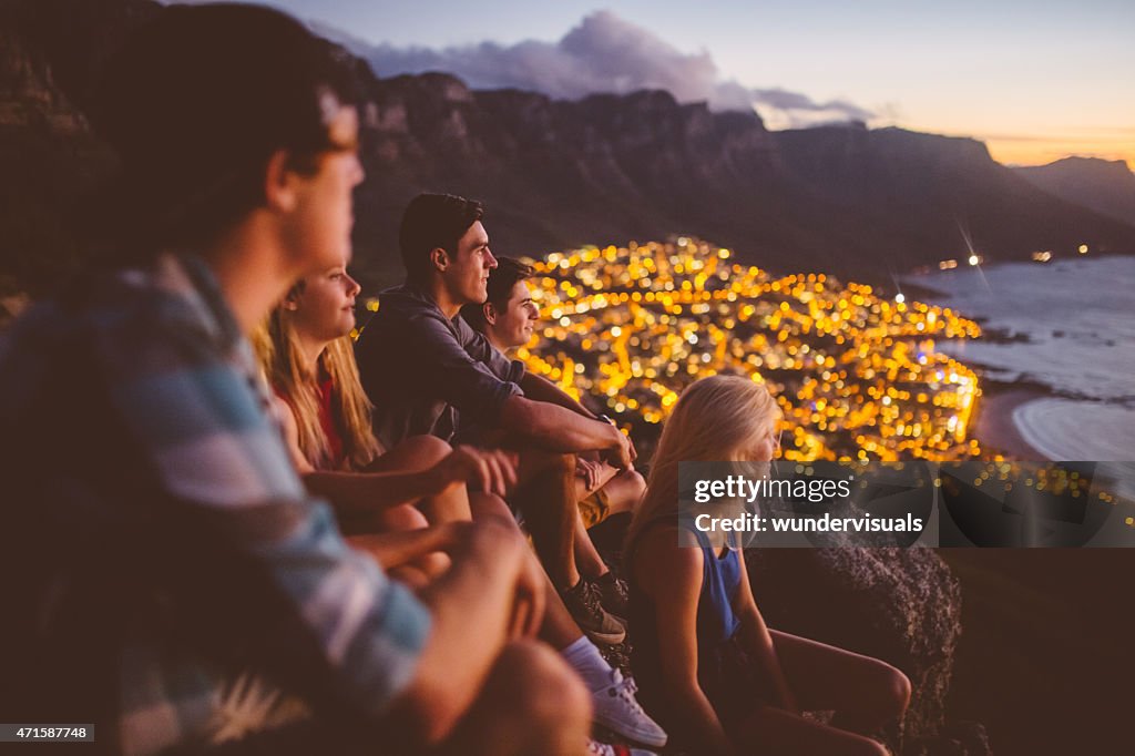Teens sitting on mountain at sunset with city lights beyond