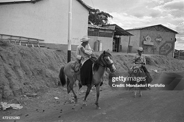 "Two men having fun riding a horse and a donkey. Behind them, the advertising of Coca Cola. Caracas, January 1958 "