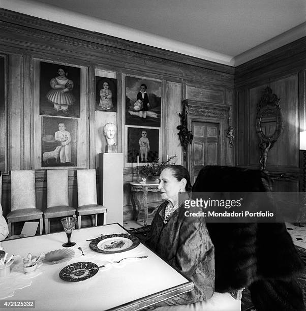 "Helena Rubinstein sits on a chair covered with a fur, at the head of a laid table, into the lavishly dining room of her house; at the walls, paneled...