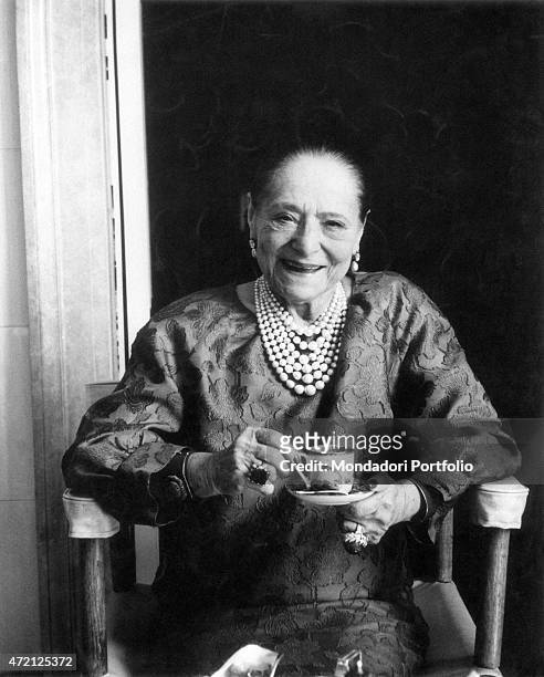 "Portrait of a smiling Helena Rubinstein, seated on a chair with a china cup in hand; the famous Polish born American entrepreneur of cosmetics...