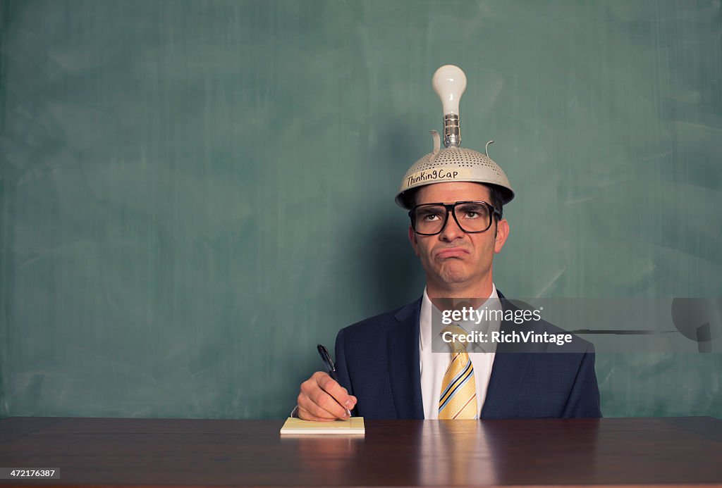 Confused Businessman Wearing Unlit Thinking Cap at Desk
