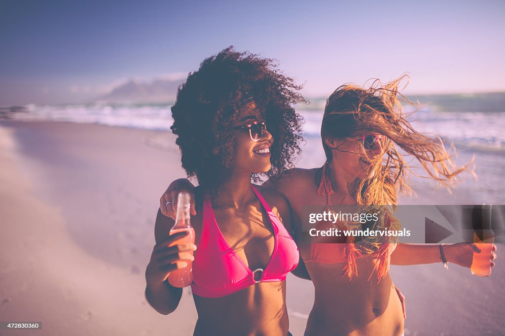 Afro niña y su amigo caminando en la playa con alcopops